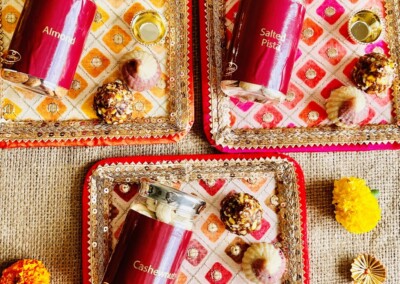 Floral Art presents Three decorated trays with jars labeled "Almond," "Salted Pista," and "Cashewnut" surrounded by sweets and floral jewelry on a woven fabric background.