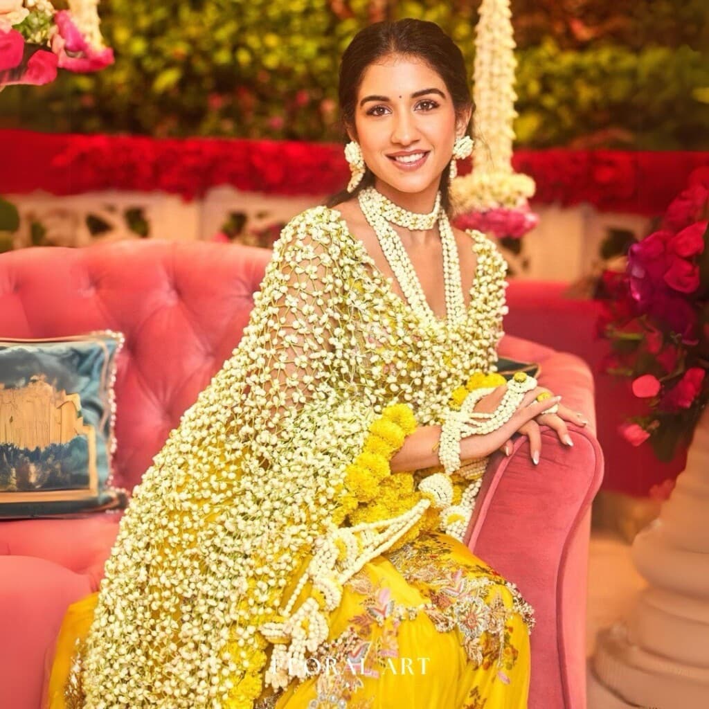 Floral Art presents A woman dressed in an elaborate, floral outfit sits on a pink couch. She is smiling and surrounded by colorful, festive decor.