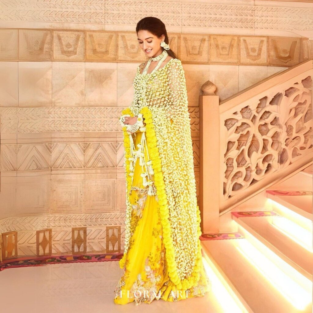 Floral Art presents A woman in a yellow traditional dress adorned with white flower garlands stands on a decorated staircase, smiling while looking down.