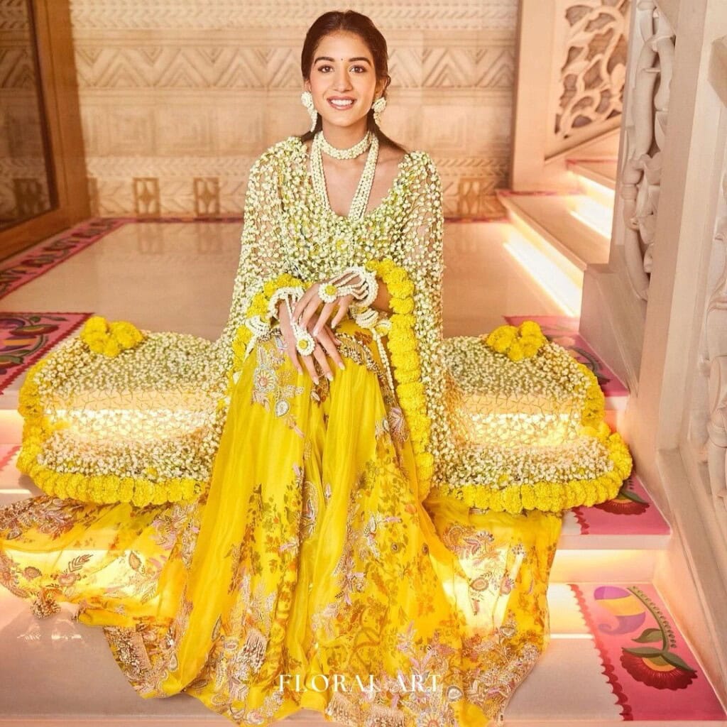 Floral Art presents A woman in a yellow and floral outfit sits on indoor stairs adorned with flowers, smiling.