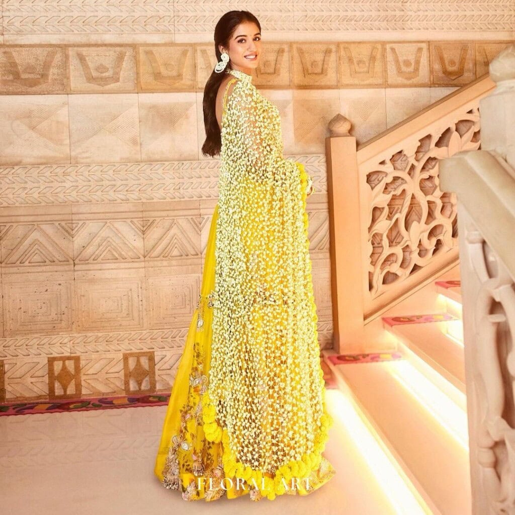 Floral Art presents A woman in a vibrant yellow traditional outfit poses on a staircase adorned with intricate designs. She smiles while turning to face the camera, holding the embellished drape of her attire.