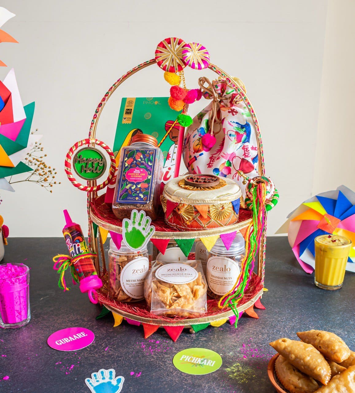 Floral Art presents A festive gift basket with snacks, drinks, and colorful decorations, surrounded by powdered colors, samosas, and paper crafts on a table.